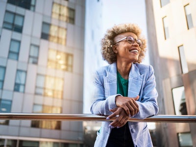 The picture shows a young woman standing on a raised platform or balcony. She is leaning casually over a shiny metal railing and smiling happily. The woman has blonde, curly hair with a fashionable undercut and wears large, round glasses. She is dressed in a light blue chequered blazer and a green T-shirt. In the background you can see modern office buildings with lots of windows. These buildings are grey and reflect the sunlight, giving the picture a dynamic and lively atmosphere. The light comes from the right-hand side and creates bright reflections on the windows and the railings. The woman is in the centre of the picture, leaning slightly to the right as she grasps the railing. Her facial expression is relaxed and content, as if she is enjoying the view and the moment. The setting suggests an urban location, probably in a city with modern architectural elements.