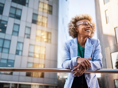 The picture shows a young woman standing on a raised platform or balcony. She is leaning casually over a shiny metal railing and smiling happily. The woman has blonde, curly hair with a fashionable undercut and wears large, round glasses. She is dressed in a light blue chequered blazer and a green T-shirt. In the background you can see modern office buildings with lots of windows. These buildings are grey and reflect the sunlight, giving the picture a dynamic and lively atmosphere. The light comes from the right-hand side and creates bright reflections on the windows and the railings. The woman is in the centre of the picture, leaning slightly to the right as she grasps the railing. Her facial expression is relaxed and content, as if she is enjoying the view and the moment. The setting suggests an urban location, probably in a city with modern architectural elements.