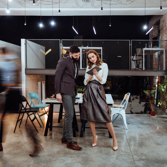 The picture shows a modern, open-plan office space where two people are standing in the center of the room. In the background on the left are cupboards containing files and a presentation board. To the right, you can see two white walls raised close together that do not reach the ceiling of the room. A railing is attached to the right-hand wall, which extends to the right-hand side of the picture and to the wall there. The wall of the railing itself is made of a grey grid and the floor is attached to a steel beam that can be seen from the side. The lattice provides a clear view of the black wall behind it. Two large desks can be seen in the center of this elevation, with two chairs on either side. On the right-hand side, against the brick wall, there is another desk with a chair and a passageway in the wall. In the foreground you can see two people walking towards the cupboards on the left. These two people are only vaguely recognizable as they are in motion and therefore blurry. In front of the railing in the center of the picture are two large tables that have been placed opposite each other and form a large group of tables. There are two chairs to the left and right of the group of tables. Three plants can be seen on the right-hand edge of the picture, their branches protruding into the picture. In the center of the picture, a younger man is standing next to a younger woman, right before the group of tables. The man has short black hair and a black beard and is wearing a black jacket with grey trousers and brown shoes. The woman has open red wavy hair and is wearing a white blouse, a grey metallic skirt and white pumps. They are both smiling while the woman has a tablet in her hand and is presenting the smart metering options from ista to the man.