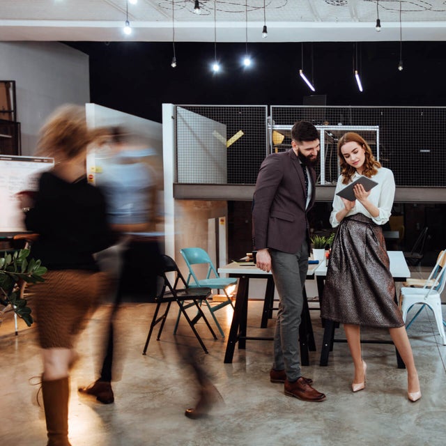 The picture shows a modern, open-plan office space where two people are standing in the center of the room. In the background on the left are cupboards containing files and a presentation board. To the right, you can see two white walls raised close together that do not reach the ceiling of the room. A railing is attached to the right-hand wall, which extends to the right-hand side of the picture and to the wall there. The wall of the railing itself is made of a grey grid and the floor is attached to a steel beam that can be seen from the side. The lattice provides a clear view of the black wall behind it. Two large desks can be seen in the center of this elevation, with two chairs on either side. On the right-hand side, against the brick wall, there is another desk with a chair and a passageway in the wall. In the foreground you can see two people walking towards the cupboards on the left. These two people are only vaguely recognizable as they are in motion and therefore blurry. In front of the railing in the center of the picture are two large tables that have been placed opposite each other and form a large group of tables. There are two chairs to the left and right of the group of tables. Three plants can be seen on the right-hand edge of the picture, their branches protruding into the picture. In the center of the picture, a younger man is standing next to a younger woman, right before the group of tables. The man has short black hair and a black beard and is wearing a black jacket with grey trousers and brown shoes. The woman has open red wavy hair and is wearing a white blouse, a grey metallic skirt and white pumps. They are both smiling while the woman has a tablet in her hand and is presenting the smart metering options from ista to the man.