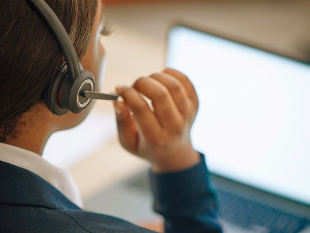 The picture shows a woman from the support center with a gray headset with a microphone sitting before an open laptop that is shining in a bright white. The picture was taken over the shoulder of the woman. The woman has her hair back and is wearing a white shirt and a dark blue jarkett and with er right hand she is adjusting the microphone of her headset.