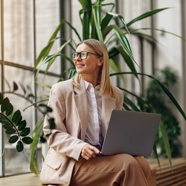 The image features a professional woman seated in a modern office setting, illuminated by natural light from large windows. She is wearing a beige blazer over a white blouse and matching beige pants. The woman has shoulder-length blonde hair and is wearing glasses. She is holding a laptop on her lap and looking outside with a pleasant and focused expression, because the energy accounting from ista is making everything easier. The background includes several lush green plants, adding a touch of nature to the workspace.