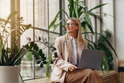 The image features a professional woman seated in a modern office setting, illuminated by natural light from large windows. She is wearing a beige blazer over a white blouse and matching beige pants. The woman has shoulder-length blonde hair and is wearing glasses. She is holding a laptop on her lap and looking outside with a pleasant and focused expression, because the energy accounting from ista is making everything easier. The background includes several lush green plants, adding a touch of nature to the workspace.