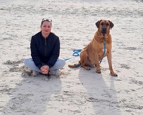 Eine Frau sitzt in einem Schneidersitz auf einem sandigen Strand neben einem großen braunen Hund. Beide schauen in die Kamera. Der Hund ist an einer blauen Leine befestigt.