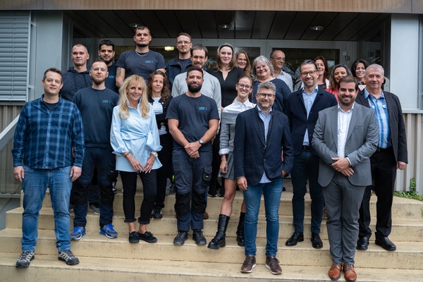 Ein Gruppenfoto von etwa 20 Personen, die auf einer Außentreppe vor einem Gebäudeeingang stehen. Die Gruppe besteht aus Männern und Frauen unterschiedlichen Alters, einige sind lässig gekleidet, andere tragen Businesskleidung. Die Personen lächeln und schauen in die Kamera. Vorne stehen zwei Männer in Anzügen, einer in einem dunkelgrauen und der andere in einem hellgrauen Anzug, sowie eine Frau in einer hellblauen Bluse. Im Hintergrund ist ein Teil der Gebäudefassade zu sehen.