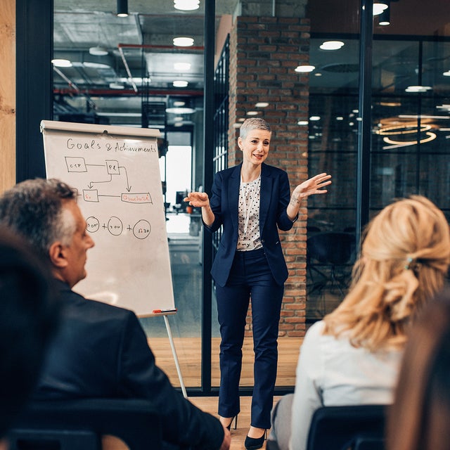 The picture shows a woman in a blue business outfit next to a board. In the lower part of the picture, several employees can be seen listening attentively to her. This scene conveys a professional and concentrated working atmosphere.