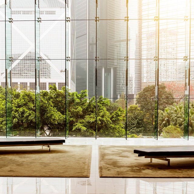 The picture shows a modern lobby or waiting area with an impressive glass front that offers a view of an urban landscape. In the foreground of the picture are two plain black benches standing on a large beige carpet. The benches are arranged symmetrically to each other, one on the left and one on the right, leaving an empty space between them. The floor is made of polished white tiles that reflect the surroundings and the incoming light, creating a bright and inviting impression. The glass wall in the background consists of several tall, narrow panels that reach up to the ceiling and are connected by metal struts. Green, lush vegetation can be seen through the glass wall, contrasting with the tall, modern office buildings in the background. The buildings have different architectural styles and reflect the light in different ways, giving the scene depth and dynamism. The left side shows a striking building with a characteristic diagonal structure, while the right side shows several high-rise buildings with glass facades. The sun shines into the picture from the top right, bathing the scene in a warm, golden light.