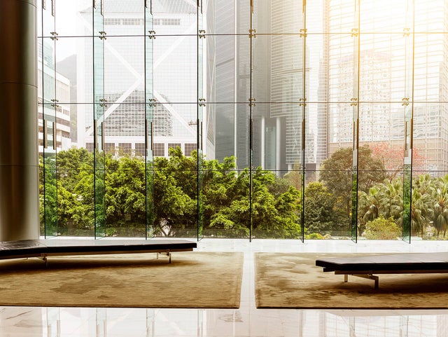 The picture shows a modern lobby or waiting area with an impressive glass front that offers a view of an urban landscape. In the foreground of the picture are two plain black benches standing on a large beige carpet. The benches are arranged symmetrically to each other, one on the left and one on the right, leaving an empty space between them. The floor is made of polished white tiles that reflect the surroundings and the incoming light, creating a bright and inviting impression. The glass wall in the background consists of several tall, narrow panels that reach up to the ceiling and are connected by metal struts. Green, lush vegetation can be seen through the glass wall, contrasting with the tall, modern office buildings in the background. The buildings have different architectural styles and reflect the light in different ways, giving the scene depth and dynamism. The left side shows a striking building with a characteristic diagonal structure, while the right side shows several high-rise buildings with glass facades. The sun shines into the picture from the top right, bathing the scene in a warm, golden light.