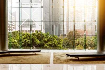 The picture shows a modern lobby or waiting area with an impressive glass front that offers a view of an urban landscape. In the foreground of the picture are two plain black benches standing on a large beige carpet. The benches are arranged symmetrically to each other, one on the left and one on the right, leaving an empty space between them. The floor is made of polished white tiles that reflect the surroundings and the incoming light, creating a bright and inviting impression. The glass wall in the background consists of several tall, narrow panels that reach up to the ceiling and are connected by metal struts. Green, lush vegetation can be seen through the glass wall, contrasting with the tall, modern office buildings in the background. The buildings have different architectural styles and reflect the light in different ways, giving the scene depth and dynamism. The left side shows a striking building with a characteristic diagonal structure, while the right side shows several high-rise buildings with glass facades. The sun shines into the picture from the top right, bathing the scene in a warm, golden light.