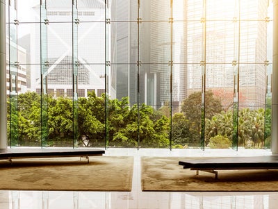 The picture shows a modern lobby or waiting area with an impressive glass front that offers a view of an urban landscape. In the foreground of the picture are two plain black benches standing on a large beige carpet. The benches are arranged symmetrically to each other, one on the left and one on the right, leaving an empty space between them. The floor is made of polished white tiles that reflect the surroundings and the incoming light, creating a bright and inviting impression. The glass wall in the background consists of several tall, narrow panels that reach up to the ceiling and are connected by metal struts. Green, lush vegetation can be seen through the glass wall, contrasting with the tall, modern office buildings in the background. The buildings have different architectural styles and reflect the light in different ways, giving the scene depth and dynamism. The left side shows a striking building with a characteristic diagonal structure, while the right side shows several high-rise buildings with glass facades. The sun shines into the picture from the top right, bathing the scene in a warm, golden light.