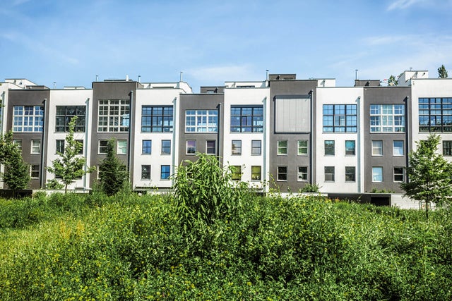 The picture shows a row of narrow but tall terraced houses. The terraced houses have alternating grey and white façades. On the left you can see a house with a white façade and on the far right a house with a grey façade. They are also always built at different heights, so that there are never two houses of the same height next to each other. All the houses have a large square window front in the upper half, consisting of 30 small square windows. The window front of the fourth house from the right is the only one covered by a blind. All of the houses have four individual windows under the fronts, with two on the left and two on the right below each other. Above the houses you can see the almost completely cloudless blue sky. In the foreground in front of the houses you can see a lawn with many bushes and a total of four trees, three of which can be seen on the left at the edge of the picture and one tree on the right.