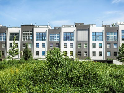 The picture shows a row of narrow but tall terraced houses. The terraced houses have alternating grey and white façades. On the left you can see a house with a white façade and on the far right a house with a grey façade. They are also always built at different heights, so that there are never two houses of the same height next to each other. All the houses have a large square window front in the upper half, consisting of 30 small square windows. The window front of the fourth house from the right is the only one covered by a blind. All of the houses have four individual windows under the fronts, with two on the left and two on the right below each other. Above the houses you can see the almost completely cloudless blue sky. In the foreground in front of the houses you can see a lawn with many bushes and a total of four trees, three of which can be seen on the left at the edge of the picture and one tree on the right.