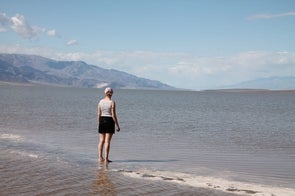 Eine Person steht mit dem Rücken zur Kamera am Rand eines Gewässers. Sie trägt ein gestreiftes Oberteil, schwarze Shorts und eine rosa Mütze. Im Hintergrund ist die Wüste und eine Gebirgskette sichtbar.