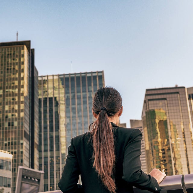 The picture shows a woman standing at a silver railing and looking at a view of many skyscrapers. She has long brown hair tied back in a ponytail and is wearing a black jacket. She is standing with her back to the viewer. The skyscrapers are close together and all have either complete window fronts or many rows of windows stretching through the entire building. The sun shines on the windows of some of the buildings, while the sun shines only partially on the other buildings, because some of the other skyscrapers are casting shadows on them.