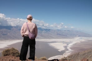 Eine Person steht mit dem Rücken zur Kamera auf einem Aussichtspunkt mit Blick auf eine weite Wüstenlandschaft mit Salzflächen und Bergen im Hintergrund. Sie trägt eine rosa Mütze, einen rosa Pullover, eine schwarze Hose und eine rosa Umhängetasche.