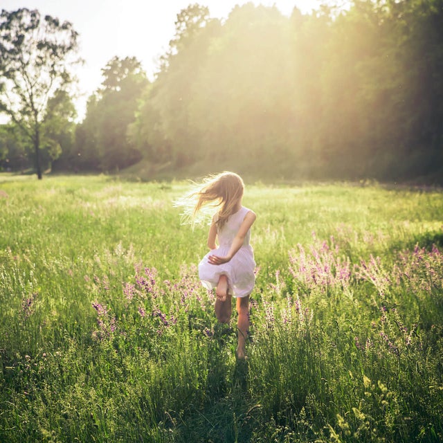 The picture shows a green, wide and tall meadow with large green trees on the edge of the horizon in the upper third of the picture and a girl running through the meadow. From the upper edge in the centre, bright yellow rays of sunlight stretch across the middle of the picture and bathe it in warm light. In the centre of the picture, a little girl in a knee-length, sleeveless white dress is walking across the meadow with her back to the viewer. Her long blonde hair blows away from her body to the left as she moves. In addition, as she runs, her left arm is loosely behind her back and her left foot is bent upwards so that the entire underside of her foot can be seen at the end of her dress.