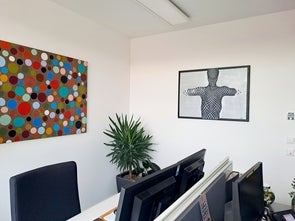 Ein modernes Büro mit zwei Gemälden an der Wand. Links ist ein buntes Gemälde mit vielen verschiedenfarbigen Punkten, und rechts ein Schwarz-Weiß-Bild einer stilisierten menschlichen Figur. Im Vordergrund befinden sich Bürotische und ein grüner Zimmerpflanze.