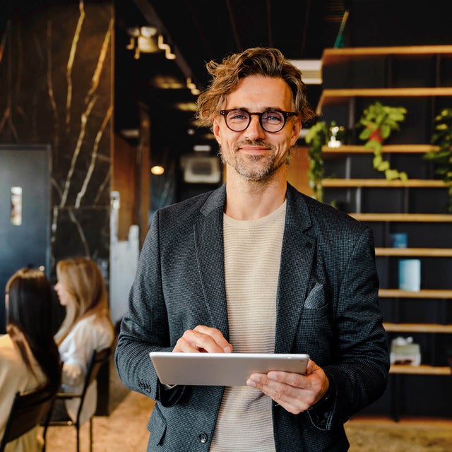 The picture shows a smiling man holding a tablet on which ista's Energy Management Minute View is displayed. The man has brown hair and is wearing black glasses. He is wearing a beige and grey striped shirt and a blue blazer over it. He is standing in the centre of the picture and looking directly into the camera. A modern office can be seen in the background. On the left-hand side of the picture is a wooden shelf in front of a black wall, decorated with long hanging plants and various objects that give the impression of awards. On the right of the picture, two women are sitting next to each other at a white work table covered with various work items. The woman in front, whose long black hair and beige loose top are visible, rests her arms on the table and has turned her head towards her colleague, so that only the back of her head is visible. The woman next to her has long blonde hair and is wearing a light blue shirt. Her side profile is recognisable. The entire background is blurred so that the focus is on the smiling man with the tablet.