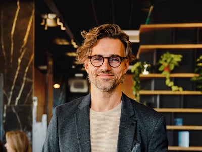 The picture shows a smiling man holding a tablet on which ista's Energy Management Minute View is displayed. The man has brown hair and is wearing black glasses. He is wearing a beige and grey striped shirt and a blue blazer over it. He is standing in the centre of the picture and looking directly into the camera. A modern office can be seen in the background. On the left-hand side of the picture is a wooden shelf in front of a black wall, decorated with long hanging plants and various objects that give the impression of awards. On the right of the picture, two women are sitting next to each other at a white work table covered with various work items. The woman in front, whose long black hair and beige loose top are visible, rests her arms on the table and has turned her head towards her colleague, so that only the back of her head is visible. The woman next to her has long blonde hair and is wearing a light blue shirt. Her side profile is recognisable. The entire background is blurred so that the focus is on the smiling man with the tablet.