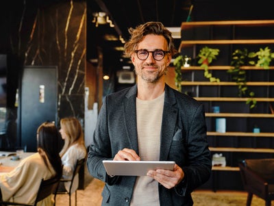 The picture shows a smiling man holding a tablet on which ista's Energy Management Minute View is displayed. The man has brown hair and is wearing black glasses. He is wearing a beige and grey striped shirt and a blue blazer over it. He is standing in the centre of the picture and looking directly into the camera. A modern office can be seen in the background. On the left-hand side of the picture is a wooden shelf in front of a black wall, decorated with long hanging plants and various objects that give the impression of awards. On the right of the picture, two women are sitting next to each other at a white work table covered with various work items. The woman in front, whose long black hair and beige loose top are visible, rests her arms on the table and has turned her head towards her colleague, so that only the back of her head is visible. The woman next to her has long blonde hair and is wearing a light blue shirt. Her side profile is recognisable. The entire background is blurred so that the focus is on the smiling man with the tablet.