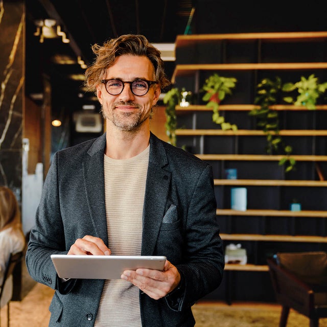 The picture shows a smiling man holding a tablet on which ista's Energy Management Minute View is displayed. The man has brown hair and is wearing black glasses. He is wearing a beige and grey striped shirt and a blue blazer over it. He is standing in the centre of the picture and looking directly into the camera. A modern office can be seen in the background. On the left-hand side of the picture is a wooden shelf in front of a black wall, decorated with long hanging plants and various objects that give the impression of awards. On the right of the picture, two women are sitting next to each other at a white work table covered with various work items. The woman in front, whose long black hair and beige loose top are visible, rests her arms on the table and has turned her head towards her colleague, so that only the back of her head is visible. The woman next to her has long blonde hair and is wearing a light blue shirt. Her side profile is recognisable. The entire background is blurred so that the focus is on the smiling man with the tablet.