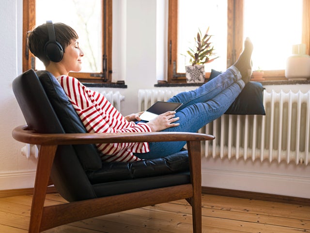 The picture shows a woman sitting comfortably on an armchair with wooden armrests and a black leather seat in front of a window. She has short brown hair and is wearing a white and red striped jumper and blue jeans. She is holding a tablet on her lap and listening to music through over-ear headphones. Her feet are propped up on a cushion resting on a radiator in front of the window. The floor of the room is made of light-coloured wood. On the windowsill, there is a flower pot with a green plant on the left and an empty white vase on the right.