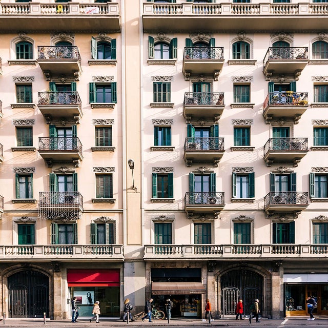 The image depicts a bustling urban street scene featuring a classic European-style apartment building. The building's facade is characterized by elegant architectural details, including wrought iron balconies, green shutters, and ornate window frames. The structure stands four stories tall, with retail shops on the ground floor, marked by large display windows and signage. Pedestrians are seen walking along the sidewalk, some engaged in conversation, while others appear to be in transit. The overall atmosphere is vibrant, reflecting a lively city environment.