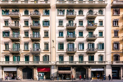 The image depicts a bustling urban street scene featuring a classic European-style apartment building. The building's facade is characterized by elegant architectural details, including wrought iron balconies, green shutters, and ornate window frames. The structure stands four stories tall, with retail shops on the ground floor, marked by large display windows and signage. Pedestrians are seen walking along the sidewalk, some engaged in conversation, while others appear to be in transit. The overall atmosphere is vibrant, reflecting a lively city environment.