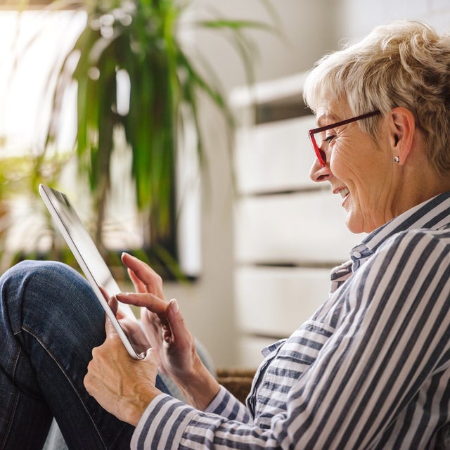 The picture shows a relaxed scene in which an elderly woman is sitting comfortably on the floor. She is turned sideways to the camera and leaning slightly backwards against a white cupboard. Her right arm is bent while she holds the tablet in both hands and touches the screen with the fingers of her right hand. The woman is wearing glasses with red frames and a blue and white striped shirt. Her hair is short and blonde and she smiles as she looks at the tablet. There are several green plants in pots in the background. The largest plant is at the top left of the picture, right next to a window. This window allows warm, natural light to stream into the room, creating a cosy and bright atmosphere. Further left in the picture, closer to the foreground, a smaller plant can be seen, which is slightly out of focus and contributes to the warm atmosphere. To the right of the woman, in the background, other blurred plants can be seen, giving the room a fresh and lively look. These plants are at different heights and distances from the camera, adding depth to the image. The woman is sitting in a slightly bent position, with her legs drawn up and crossed. The overall effect of the room is inviting and cosy, which is enhanced by the warm light and the green plants.