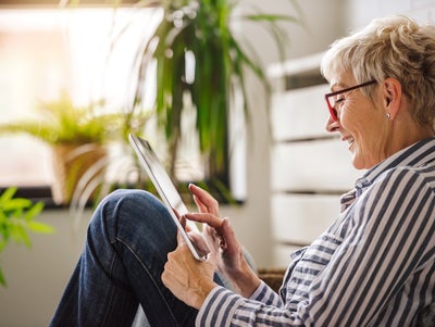 The picture shows a relaxed scene in which an elderly woman is sitting comfortably on the floor. She is turned sideways to the camera and leaning slightly backwards against a white cupboard. Her right arm is bent while she holds the tablet in both hands and touches the screen with the fingers of her right hand. The woman is wearing glasses with red frames and a blue and white striped shirt. Her hair is short and blonde and she smiles as she looks at the tablet. There are several green plants in pots in the background. The largest plant is at the top left of the picture, right next to a window. This window allows warm, natural light to stream into the room, creating a cosy and bright atmosphere. Further left in the picture, closer to the foreground, a smaller plant can be seen, which is slightly out of focus and contributes to the warm atmosphere. To the right of the woman, in the background, other blurred plants can be seen, giving the room a fresh and lively look. These plants are at different heights and distances from the camera, adding depth to the image. The woman is sitting in a slightly bent position, with her legs drawn up and crossed. The overall effect of the room is inviting and cosy, which is enhanced by the warm light and the green plants.