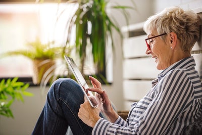 The picture shows a relaxed scene in which an elderly woman is sitting comfortably on the floor. She is turned sideways to the camera and leaning slightly backwards against a white cupboard. Her right arm is bent while she holds the tablet in both hands and touches the screen with the fingers of her right hand. The woman is wearing glasses with red frames and a blue and white striped shirt. Her hair is short and blonde and she smiles as she looks at the tablet. There are several green plants in pots in the background. The largest plant is at the top left of the picture, right next to a window. This window allows warm, natural light to stream into the room, creating a cosy and bright atmosphere. Further left in the picture, closer to the foreground, a smaller plant can be seen, which is slightly out of focus and contributes to the warm atmosphere. To the right of the woman, in the background, other blurred plants can be seen, giving the room a fresh and lively look. These plants are at different heights and distances from the camera, adding depth to the image. The woman is sitting in a slightly bent position, with her legs drawn up and crossed. The overall effect of the room is inviting and cosy, which is enhanced by the warm light and the green plants.