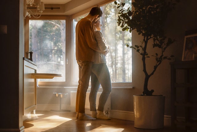 The picture shows an intimate moment in a brightly lit room. In the foreground is a couple in a loving embrace in front of a large window. The man is wearing a brown top and beige trousers, while the woman is wearing a light-coloured top and dark trousers. Both people are standing barefoot on a wooden floor, with the man standing slightly behind the woman and embracing her tenderly. The window, which takes up most of the background, allows plenty of natural light into the room and offers a view of a wooded outdoor area. The sunlight streams through the window and creates warm rays of light in the room. To the left of the picture, near the window, is a table attached to a wall. There are two small objects on the table, presumably salt and pepper shakers.  To the right of the window is a large houseplant in a white flower pot. The plant is clearly visible and lends the room a natural atmosphere. In the right-hand part of the picture, further in the background, there is a piece of wooden furniture that looks like a wine rack or a small shelf. There are a number of objects on the shelf, including a framed picture and other decorative items.