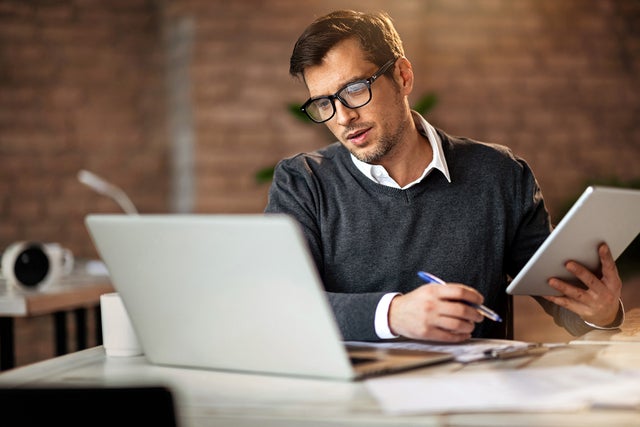 In the picture, a man wearing glasses and a gray sweater sits at a desk. On the desk, located in the foreground, are a laptop on the left and a tablet in his right hand. He is looking at the tablet while holding a pen in his left hand. In the background, there is a brick wall and blurred office items, including a mug and other objects on another desk to the left.