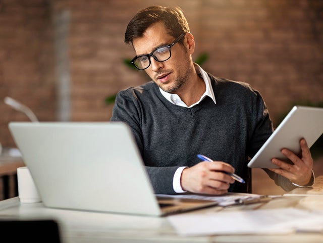 In the picture, a man wearing glasses and a gray sweater sits at a desk. On the desk, located in the foreground, are a laptop on the left and a tablet in his right hand. He is looking at the tablet while holding a pen in his left hand. In the background, there is a brick wall and blurred office items, including a mug and other objects on another desk to the left.