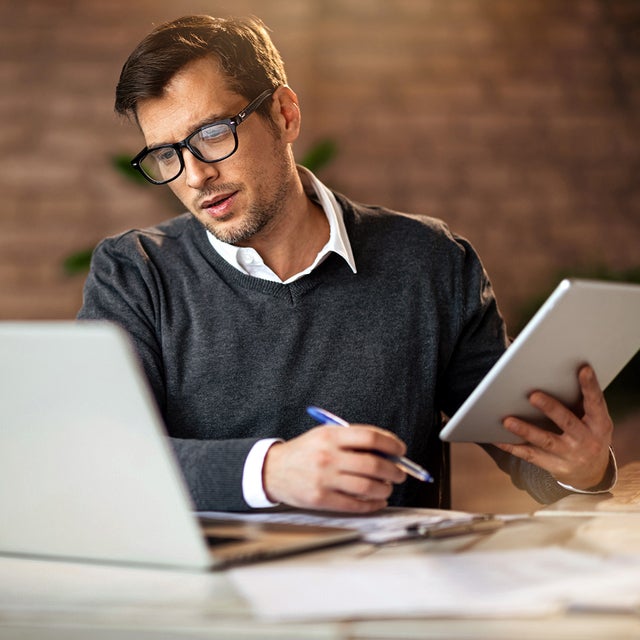 In the picture, a man wearing glasses and a gray sweater sits at a desk. On the desk, located in the foreground, are a laptop on the left and a tablet in his right hand. He is looking at the tablet while holding a pen in his left hand. In the background, there is a brick wall and blurred office items, including a mug and other objects on another desk to the left.