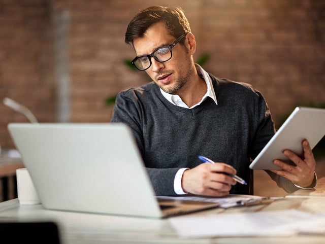 In the picture, a man wearing glasses and a gray sweater sits at a desk. On the desk, located in the foreground, are a laptop on the left and a tablet in his right hand. He is looking at the tablet while holding a pen in his left hand. In the background, there is a brick wall and blurred office items, including a mug and other objects on another desk to the left.