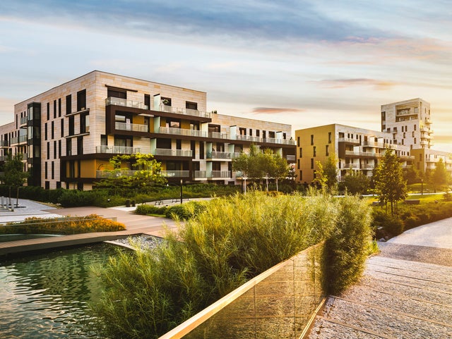 The picture shows a residential complex in the centre, stretching from the left to the right edge of the picture. The various buildings look very similar on the outside and have a white-orange brick façade. The individual flats have balconies with glass railings. The balconies are separated from each other by frosted glass walls and are furnished differently, giving each flat an individual touch. Most of the visible buildings have four floors, but vary in width so that different numbers of flats are visible on each floor. In the right quarter of the picture, in contrast to the others, another building rises up, which has the same style as the other buildings in the residential complex. A park can be seen in front of the residential complex in the lower half of the picture. Grasses and bushes surround the residential complex, accompanied by a pavement adorned with scattered trees. There are also benches and lanterns on the pavement. An artificial pond can be seen in the bottom left-hand corner, surrounded by a wooden walkway with glass railings. The sunset can be seen on the right-hand edge of the picture, giving the sky a slight colour gradient, from a reddish tone on the right to a bluish tone on the left.