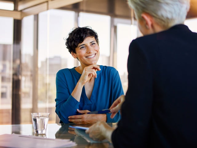 The picture shows two people sitting opposite each other at a table in a room filled with warm sunlight and with glass walls. On the left side of the table is a full glass of water and asmall pile of documents. The picture was taken from the perspective of one person, so that you only see the back of them and the other person, who is a woman, is in focus. The woman is in the center of the picture and the other person is on the right side of the picture, beeing a bit cut off. The woman is wearing a bright blue shirt, has dark wavy short hair and is smiling at the other person while holding her right hand to her chin and the other hand lying on the table touching her ellbow. The person that can only be seen from the back has a black jacket on and bright blond hair/ grey hair and a short sleek back haircut. They are explaining something to the woman while they are holding a document with their left hand and pointing with a ballpoint pen with their right hand onto something on the document.