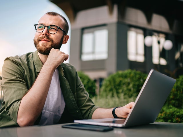 A young professional man, wearing glasses and a casual green jacket, is seated at an outdoor café with a laptop open in front of him. He appears thoughtful and focused, with his hand resting on his chin. On the table beside the laptop are a smartphone and a notepad. The background features modern office buildings and greenery. This image conveys a sense of contemplation and productivity, emphasizing the concept of consumption data monitoring in a business environment.