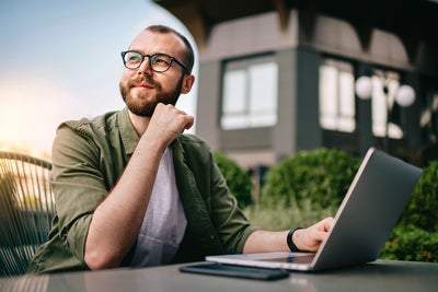 A young professional man, wearing glasses and a casual green jacket, is seated at an outdoor café with a laptop open in front of him. He appears thoughtful and focused, with his hand resting on his chin. On the table beside the laptop are a smartphone and a notepad. The background features modern office buildings and greenery. This image conveys a sense of contemplation and productivity, emphasizing the concept of consumption data monitoring in a business environment.