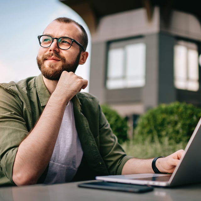 A young professional man, wearing glasses and a casual green jacket, is seated at an outdoor café with a laptop open in front of him. He appears thoughtful and focused, with his hand resting on his chin. On the table beside the laptop are a smartphone and a notepad. The background features modern office buildings and greenery. This image conveys a sense of contemplation and productivity, emphasizing the concept of consumption data monitoring in a business environment.