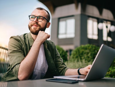 A young professional man, wearing glasses and a casual green jacket, is seated at an outdoor café with a laptop open in front of him. He appears thoughtful and focused, with his hand resting on his chin. On the table beside the laptop are a smartphone and a notepad. The background features modern office buildings and greenery. This image conveys a sense of contemplation and productivity, emphasizing the concept of consumption data monitoring in a business environment.