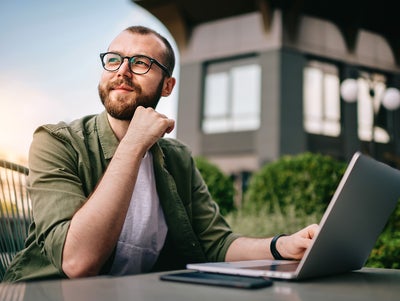 A young professional man, wearing glasses and a casual green jacket, is seated at an outdoor café with a laptop open in front of him. He appears thoughtful and focused, with his hand resting on his chin. On the table beside the laptop are a smartphone and a notepad. The background features modern office buildings and greenery. This image conveys a sense of contemplation and productivity, emphasizing the concept of consumption data monitoring in a business environment.