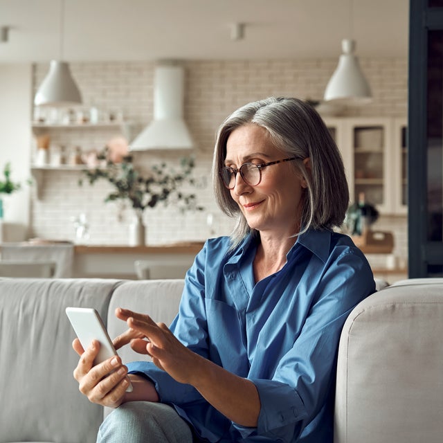 The picture shows a woman sitting peacefully on her couch and looking at her phone while smiling. background of the picture ist slightly blurred but you can still distinguish is a white modern kitchen with a white stone wall. On the stone wall are two simple shelves with food containers and to the right of them on the wall is an extractor bonnet. On the right-hand side of the picture, three wall cabinets can be seen, two of which have glass fronts. Underneath you can see a counter with two white chairs. On the counter is a vase with decorative flowers. In the foreground you can see the upper part of a light grey couch on which an elderly lady is sitting. She has a grey bob, black glasses and is wearing a blue shirt and light blue jeans. The lady is smiling at her mobile phone she is holding in her hand, with which she can easily and conveniently use ista's SmartPay option.