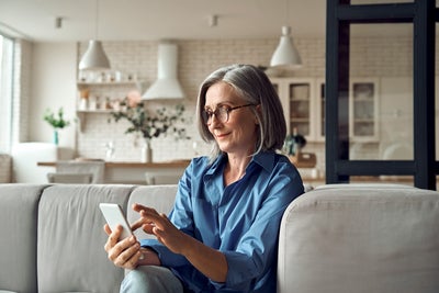 The picture shows a woman sitting peacefully on her couch and looking at her phone while smiling. background of the picture ist slightly blurred but you can still distinguish is a white modern kitchen with a white stone wall. On the stone wall are two simple shelves with food containers and to the right of them on the wall is an extractor bonnet. On the right-hand side of the picture, three wall cabinets can be seen, two of which have glass fronts. Underneath you can see a counter with two white chairs. On the counter is a vase with decorative flowers. In the foreground you can see the upper part of a light grey couch on which an elderly lady is sitting. She has a grey bob, black glasses and is wearing a blue shirt and light blue jeans. The lady is smiling at her mobile phone she is holding in her hand, with which she can easily and conveniently use ista's SmartPay option.