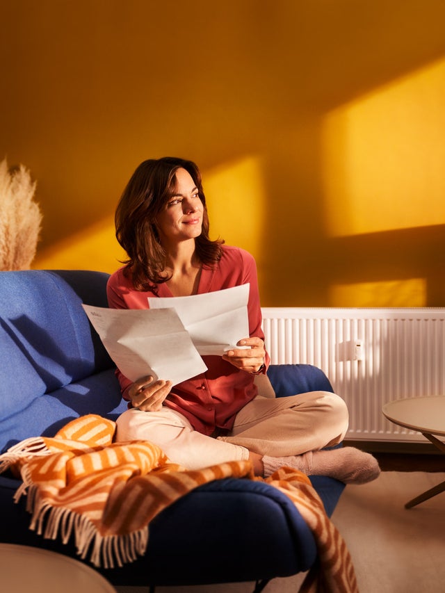 Eine brünette Frau sitzt im Schneidersitze auf ihrer blauen Couch im Wohnzimmer. In den Händen hält Sie zwei Seiten Papier, Seiten aus der Heizkostenabrechnung von ista. Sie blickt zufrieden Richtung Fenster. 