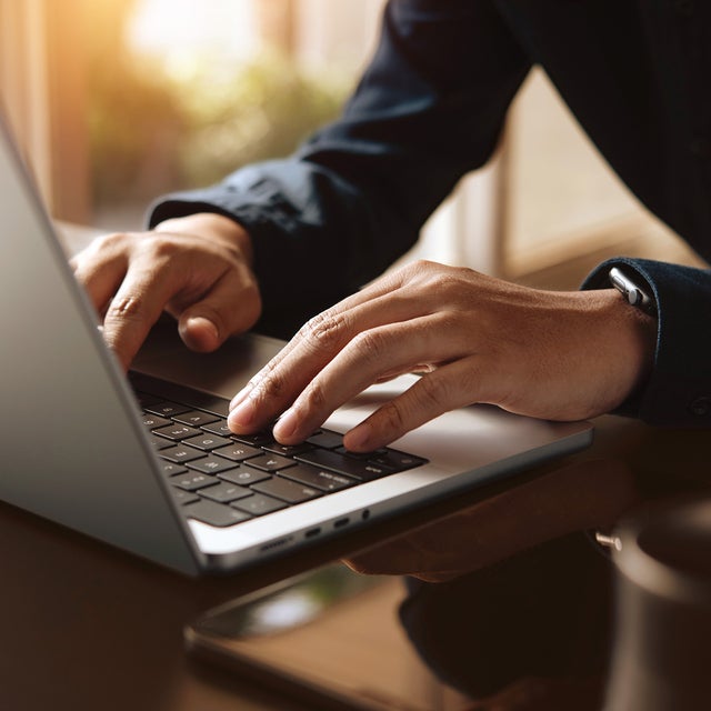 The picture shows a section of an open laptop from an angle so that only half of the keyboard is visible. A person in a blue shirt, of whom only the hands and arms can be seen, is in the right-hand half of the picture and is typing on the laptop keyboard. The hands and the laptop keyboard are in the centre of the picture and are in focus. To the right of the laptop, a smartphone and a black cup can be seen in a blur. In the background is a window that floods the room with light.