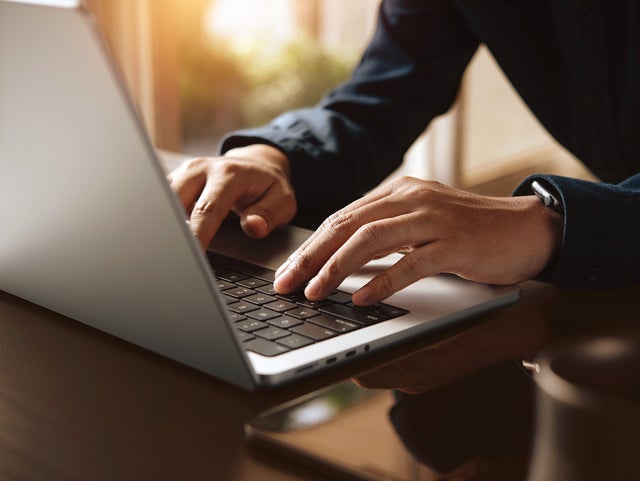 The picture shows a section of an open laptop from an angle so that only half of the keyboard is visible. A person in a blue shirt, of whom only the hands and arms can be seen, is in the right-hand half of the picture and is typing on the laptop keyboard. The hands and the laptop keyboard are in the centre of the picture and are in focus. To the right of the laptop, a smartphone and a black cup can be seen in a blur. In the background is a window that floods the room with light.