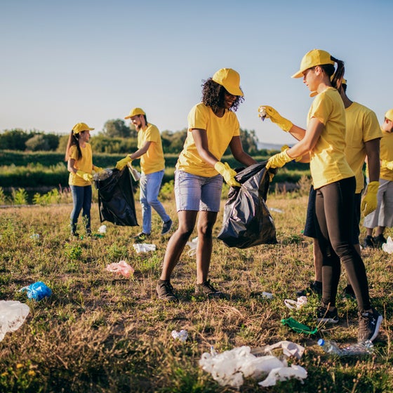 The image shows a group of people participating in a cleanup activity in an outdoor setting. They are wearing matching yellow t-shirts and hats, along with gloves, indicating they are part of an organized effort or volunteer group. The participants are picking up trash and placing it into black garbage bags. The setting appears to be a grassy field or park with some scattered litter visible on the ground. The weather is clear and sunny, and the overall atmosphere suggests teamwork and environmental stewardship.