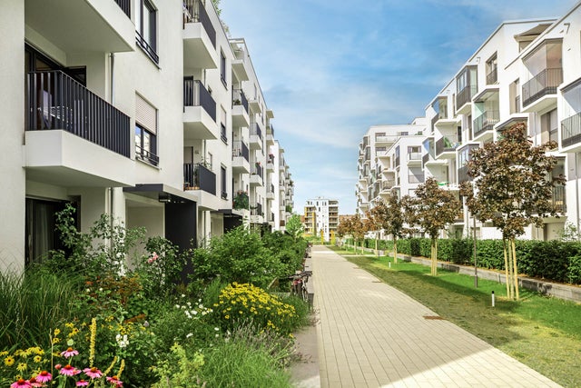 The picture shows a modern residential complex with several white, multi-storey residential buildings standing on the left and right side of a paved path. The buildings have balconies with dark railings, and some balconies are decorated with plants and furniture. A wide, paved footpath stretches between the buildings and continues into the distance. On the left side of the path are lush green areas and colourful flowerbeds that embellish the space between the buildings. On the right-hand side of the path, young trees are planted at regular intervals, forming a green avenue together with manicured lawns. Other buildings can be seen in the background, completing the urban surroundings of the residential complex. The sky is clear and blue, indicating a sunny day. Overall, the picture conveys a calm and pleasant living atmosphere in a modern, well-maintained environment.